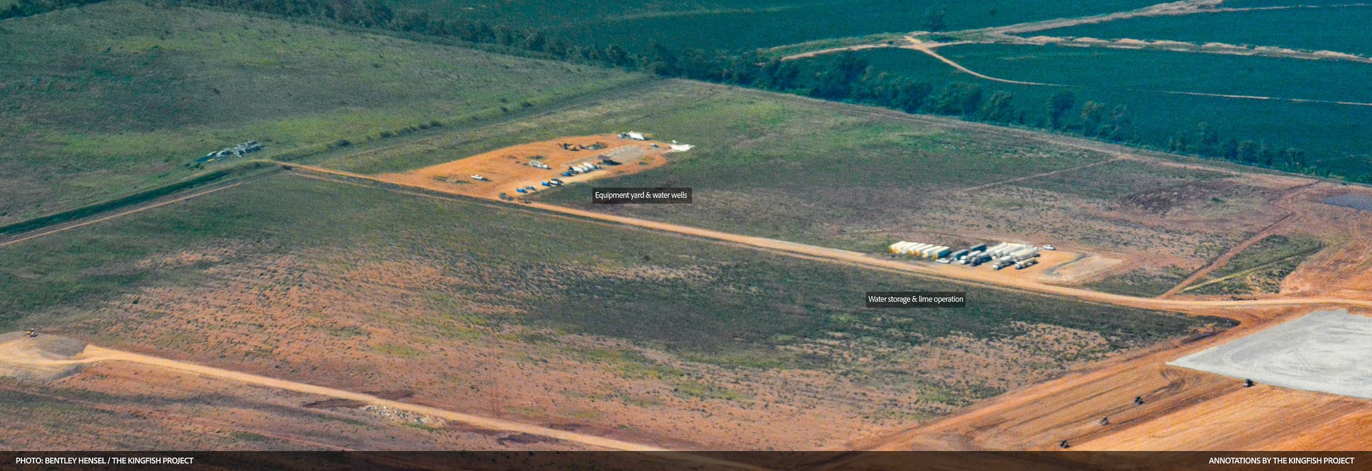 Aerial view of a large cleared construction site with two small equipment clusters along a dirt road, surrounded by green fields and tree lines.